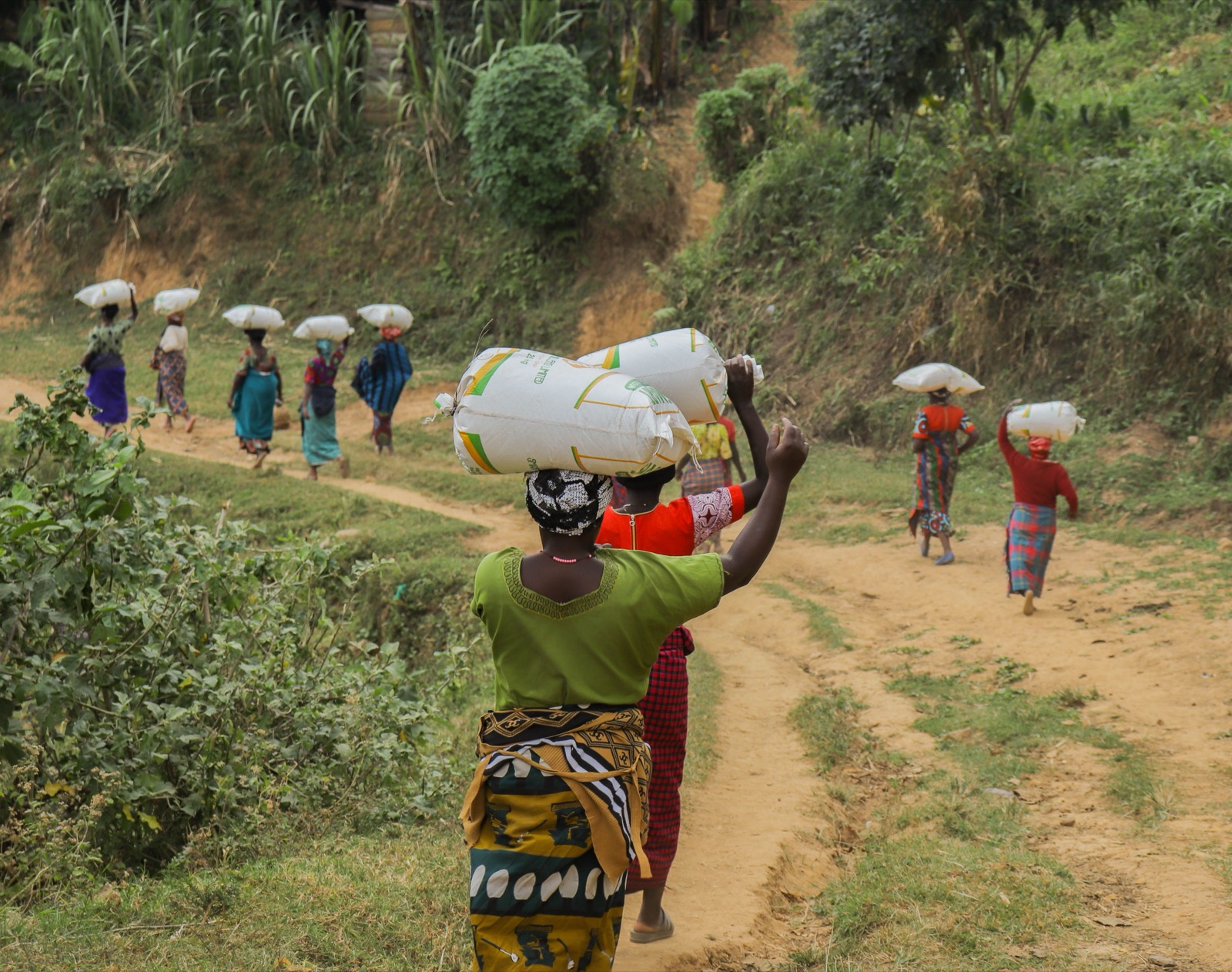 A woman carries a sack on her head along a dirt path, black and white