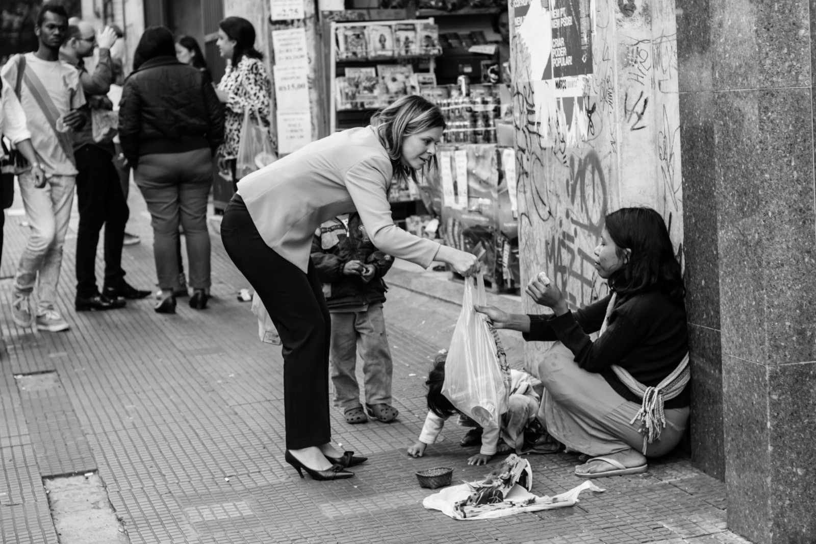 A woman hands food to a seated vendor on a sidewalk, black and white