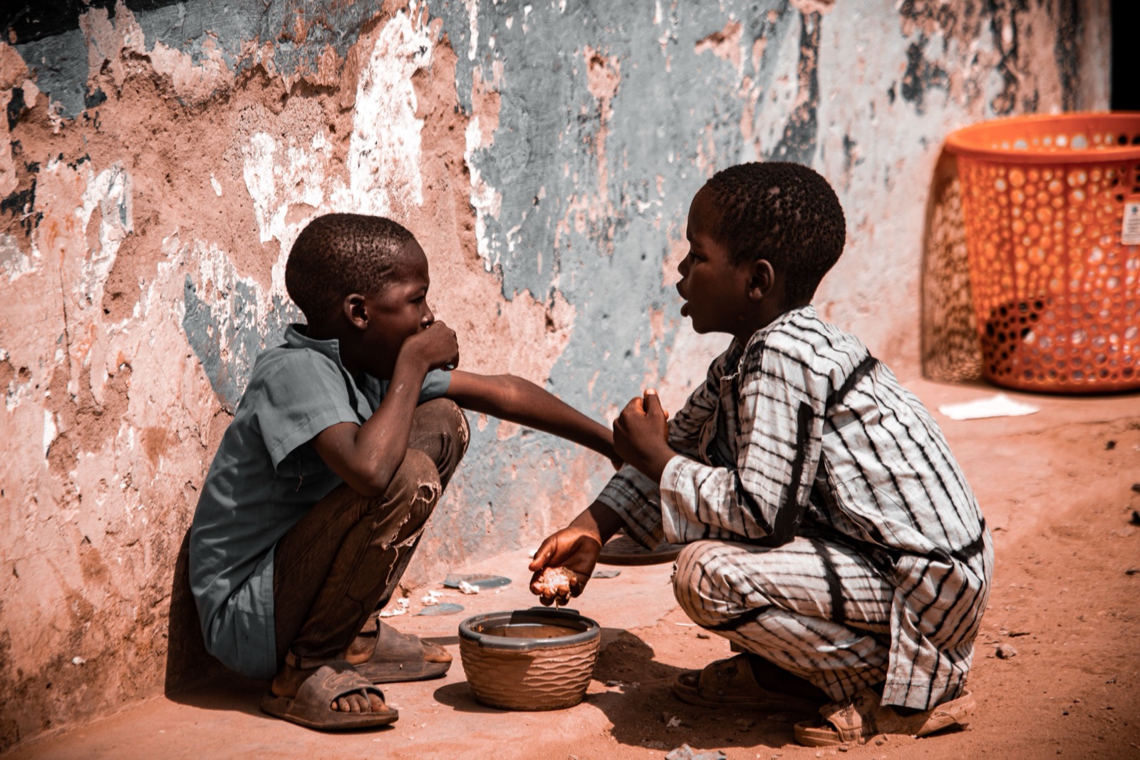 Two boys crouch and share food from a pot